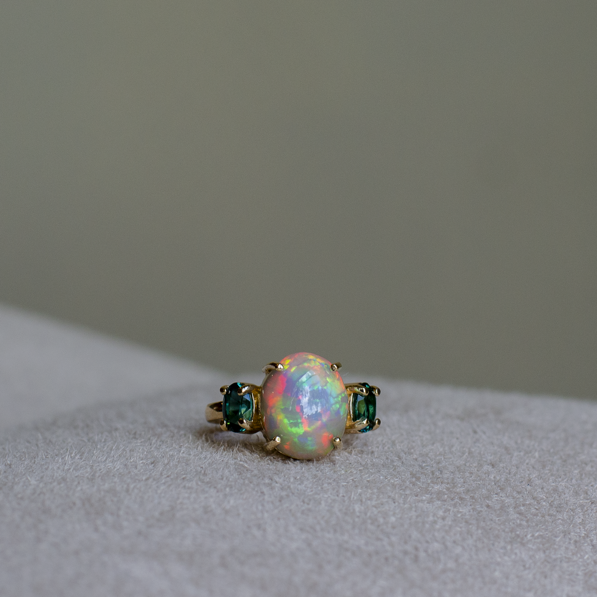 A gold ring featuring a large, round iridescent opal center stone flanked by two small oval cut blue-green indicolite tourmalines, resting on a light gray suede surface.