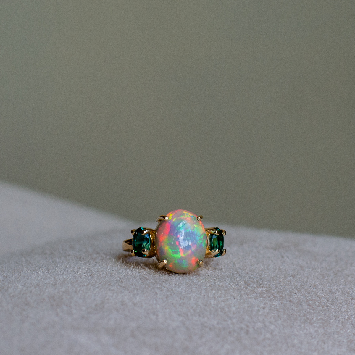 A gold ring featuring a large, round iridescent opal center stone flanked by two small oval cut blue-green indicolite tourmalines, resting on a light gray suede surface.