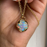 A hand holding a solid 14k yellow gold pendant with an Ethiopian opal gemstone held in a bezel setting the shape of a four leaf clover, with a gold chain.