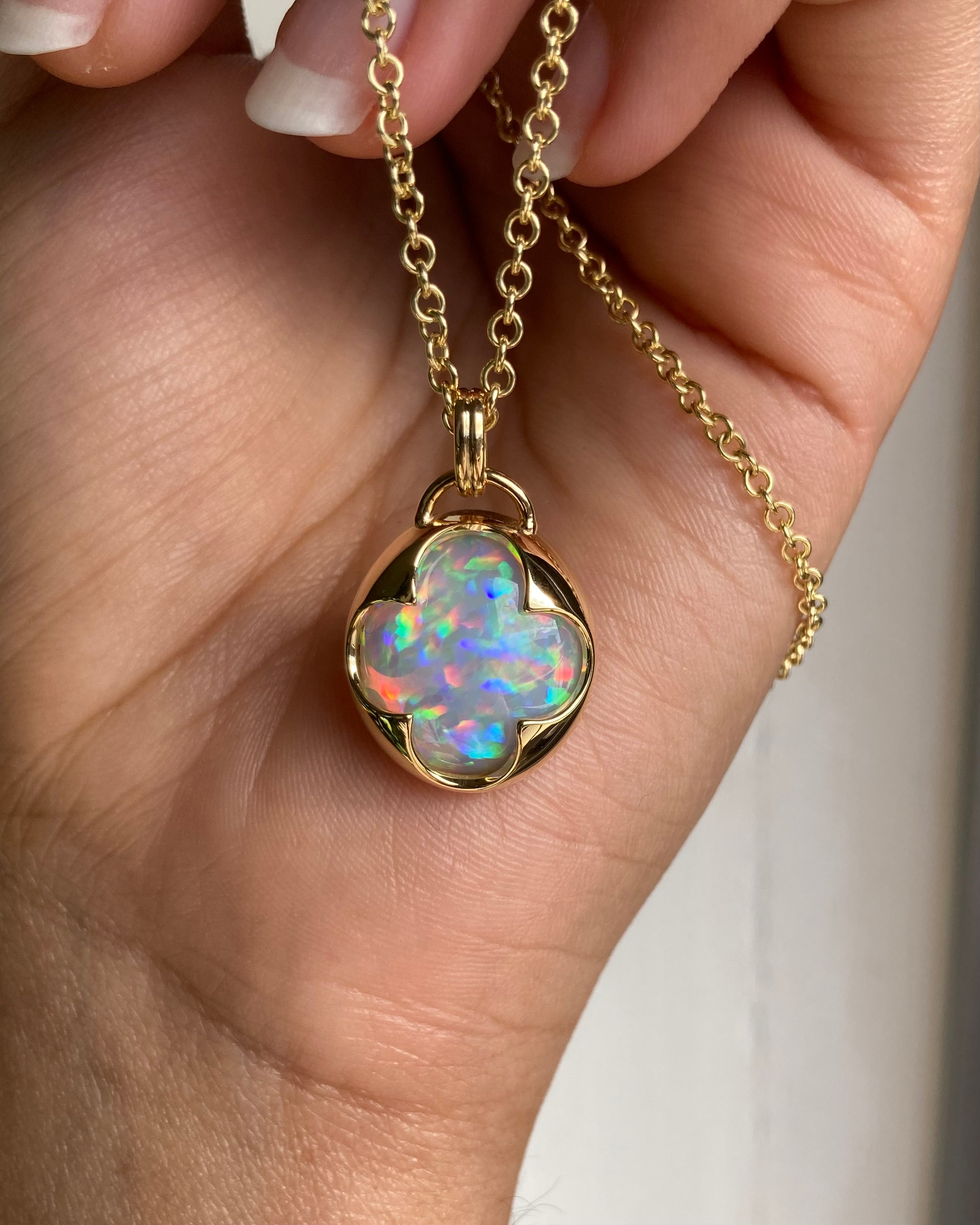 A hand holding a solid 14k yellow gold pendant with an Ethiopian opal gemstone held in a bezel setting the shape of a four leaf clover, with a gold chain.