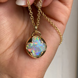 A hand holding a solid 14k yellow gold pendant with a multicolored opal gemstone held in a bezel setting the shape of a four leaf clover, with a gold chain.