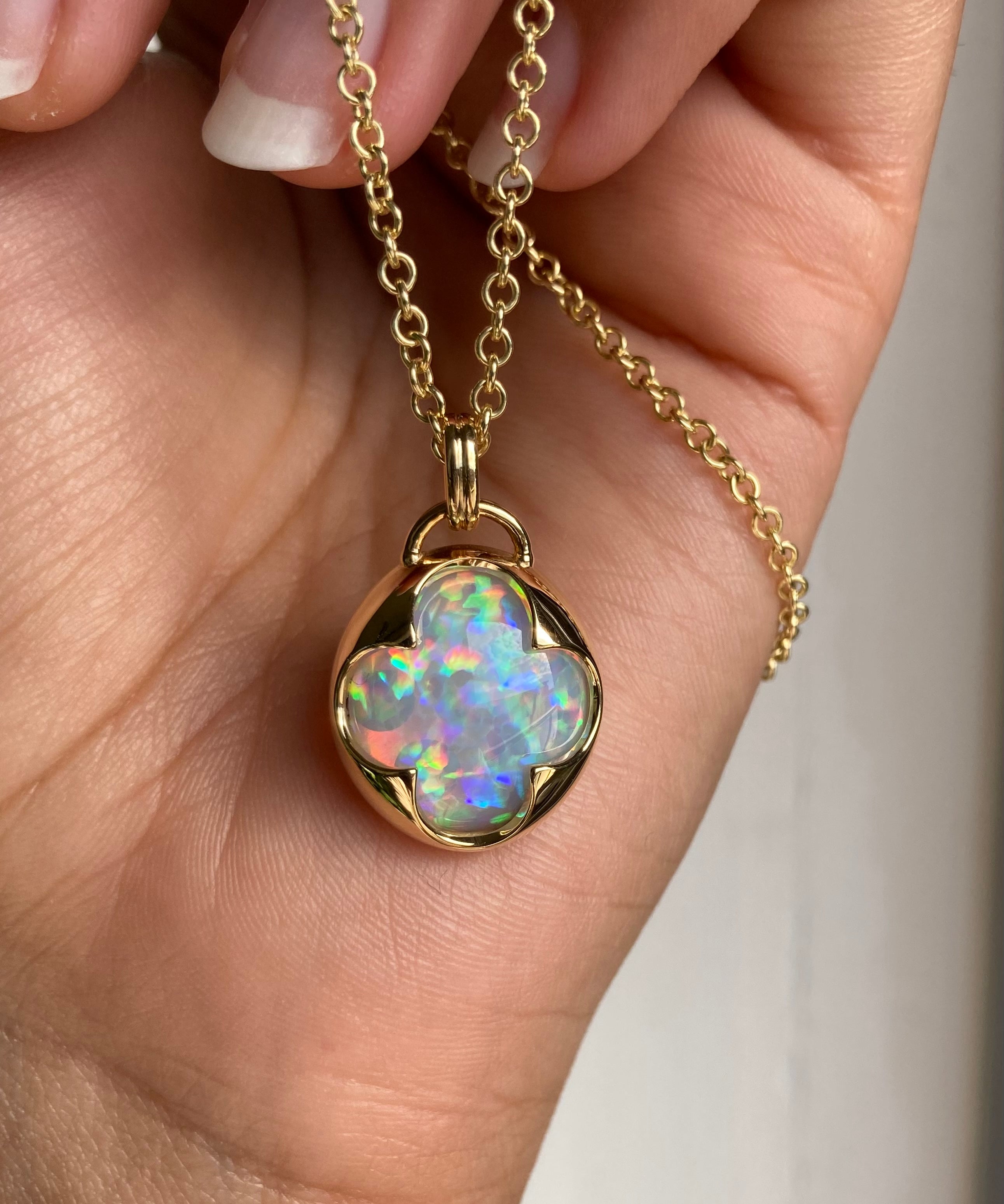 A hand holding a solid 14k yellow gold pendant with a multicolored opal gemstone held in a bezel setting the shape of a four leaf clover, with a gold chain.