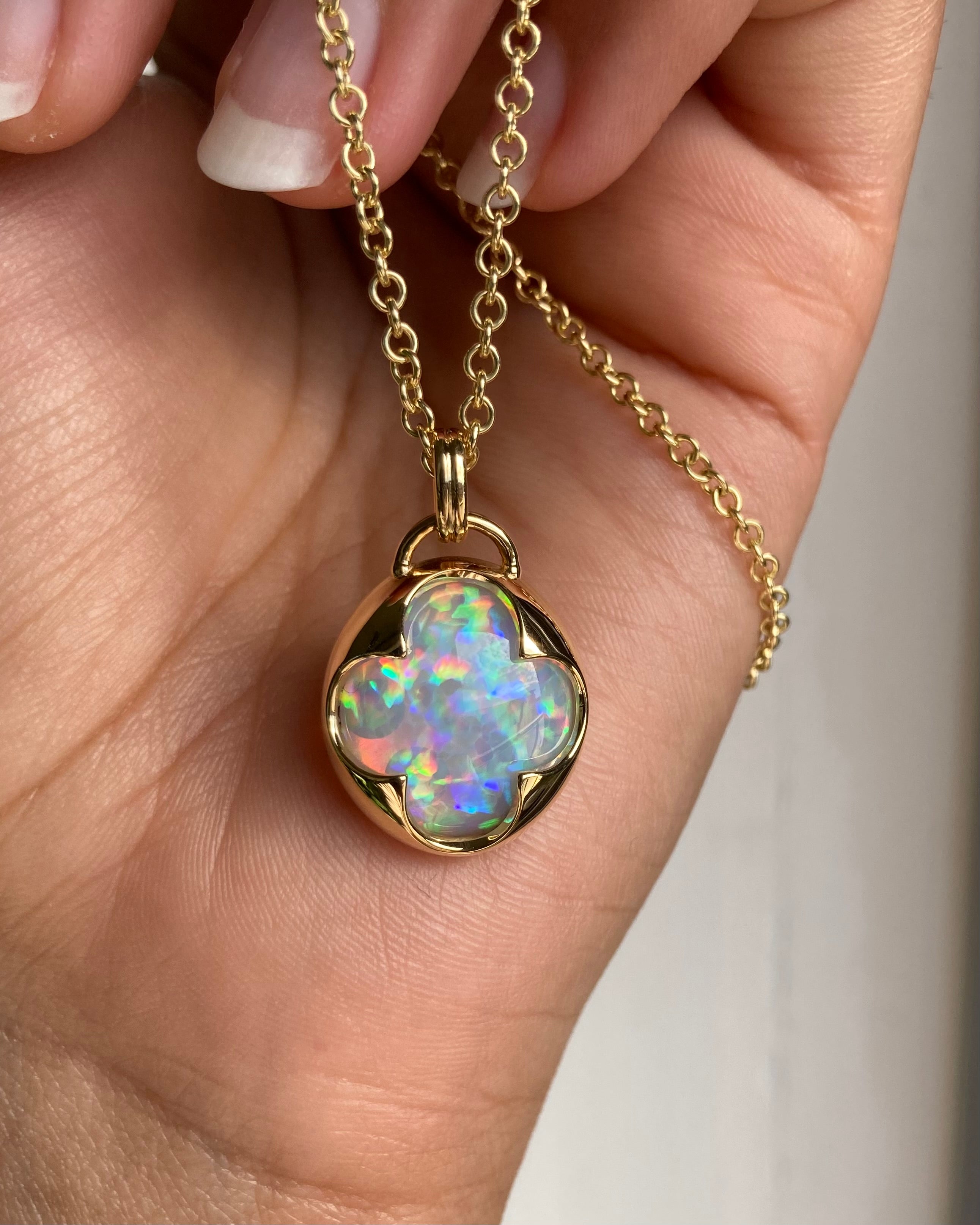 A hand holding a solid 14k yellow gold pendant with a multicolored opal gemstone held in a bezel setting the shape of a four leaf clover, with a gold chain.