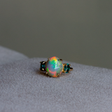 A gold ring featuring a large, round iridescent opal center stone flanked by two small oval cut blue-green indicolite tourmalines, resting on a light gray suede surface.