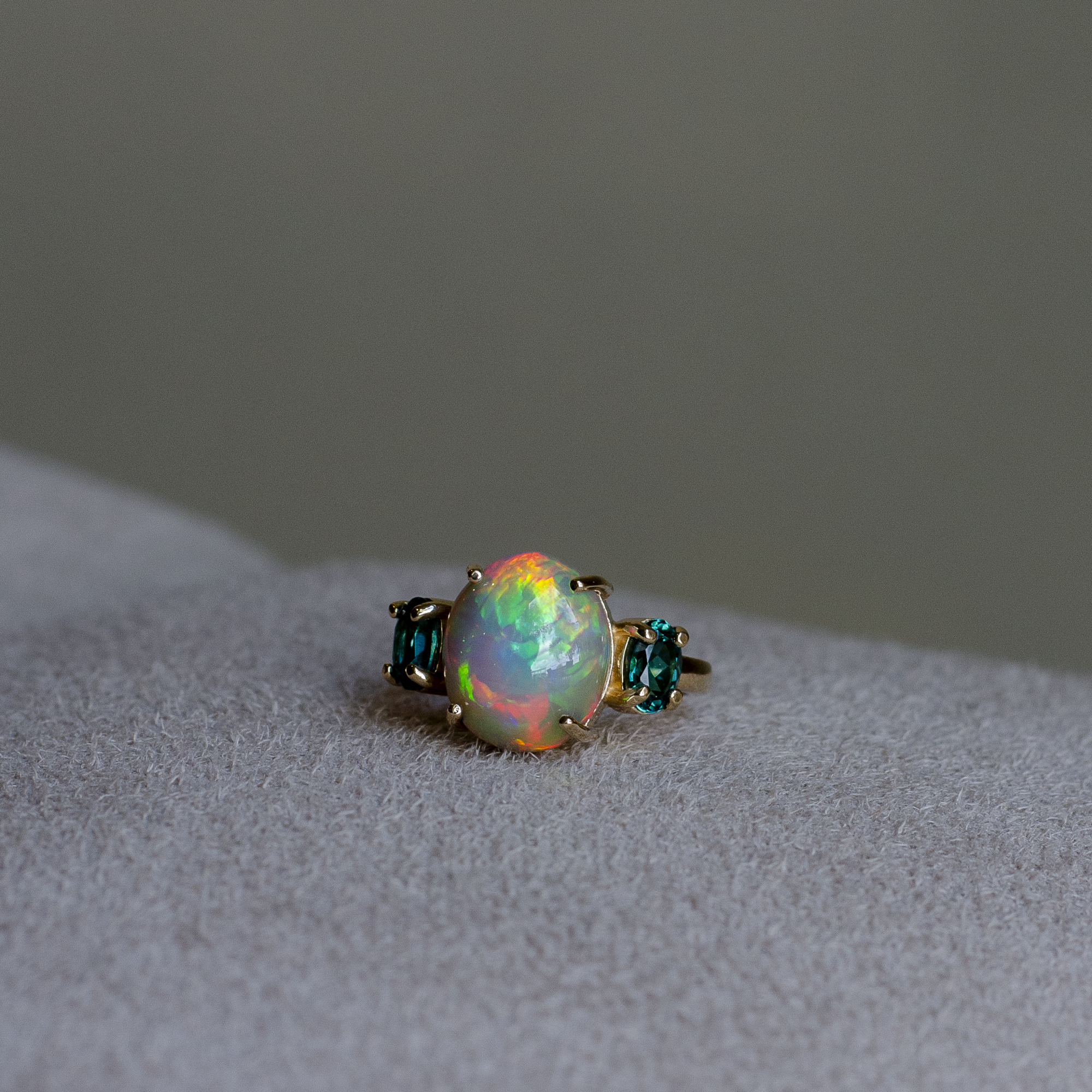 A gold ring featuring a large, round iridescent opal center stone flanked by two small oval cut blue-green indicolite tourmalines, resting on a light gray suede surface.
