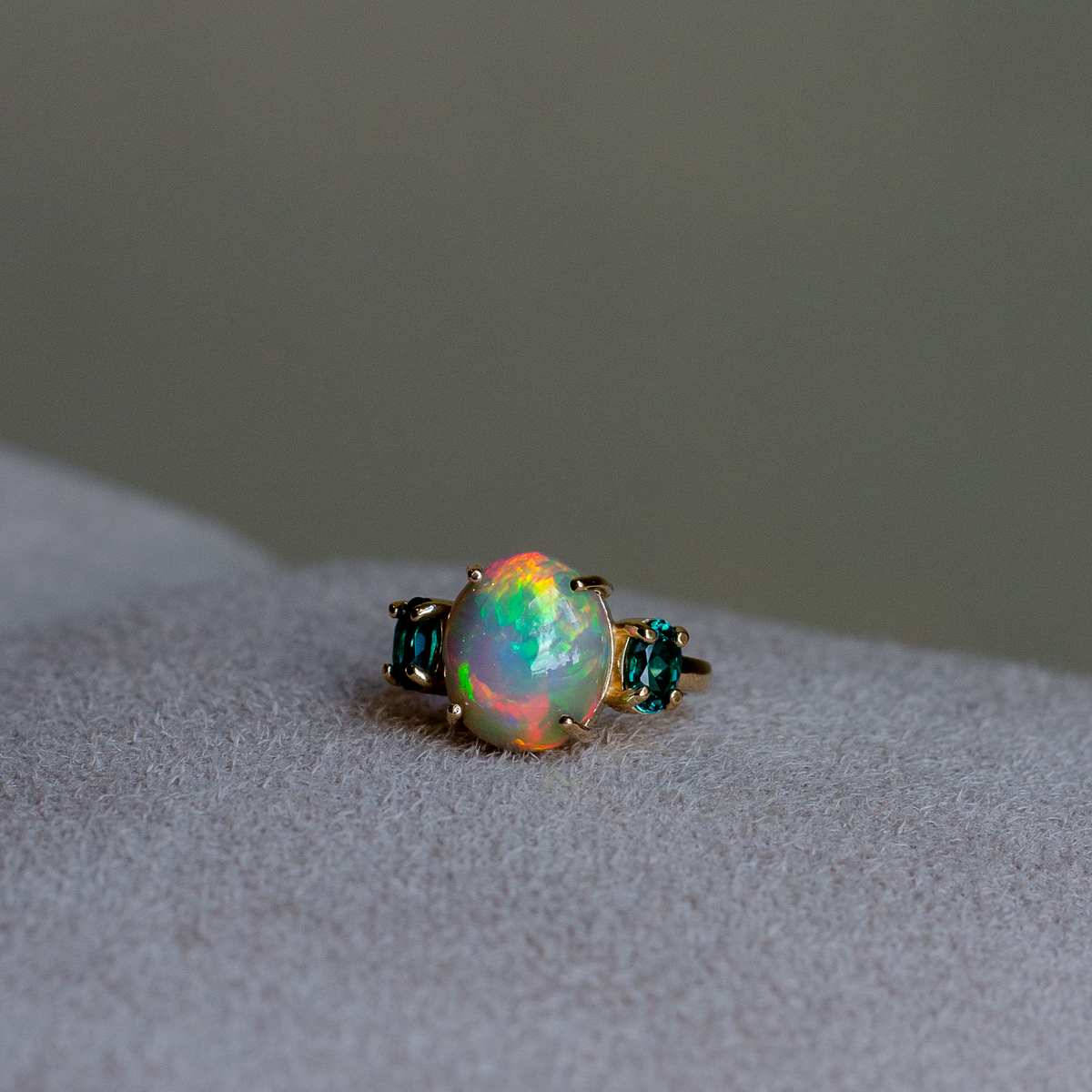 A gold ring featuring a large, round iridescent opal center stone flanked by two small oval cut blue-green indicolite tourmalines, resting on a light gray suede surface.