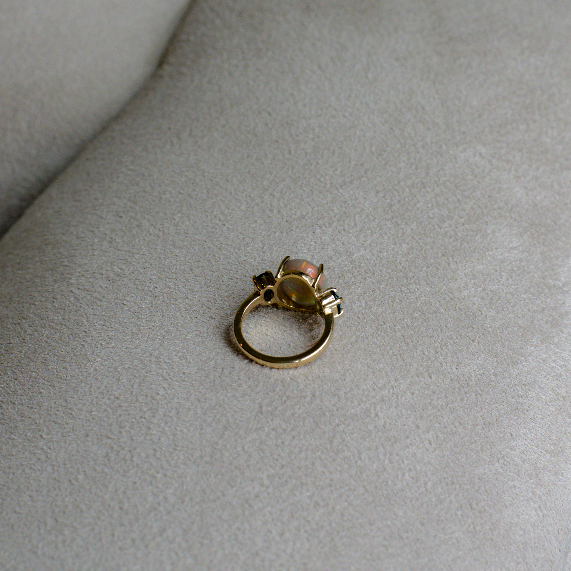Back side of a gold ring with an opal center and indicolite stones, resting on a gray suede surface.