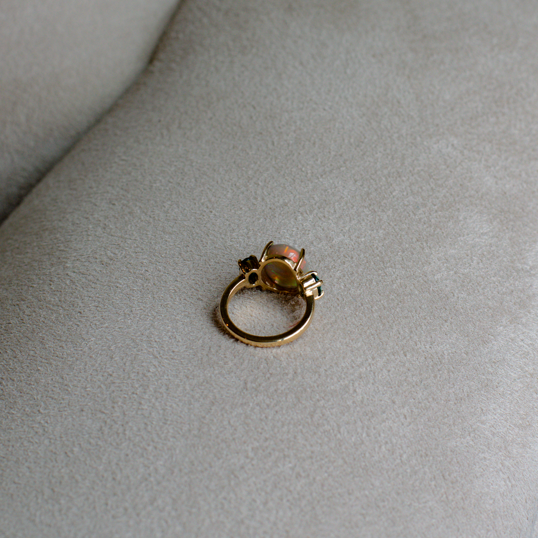Back side of a gold ring with an opal center and indicolite stones, resting on a gray suede surface.