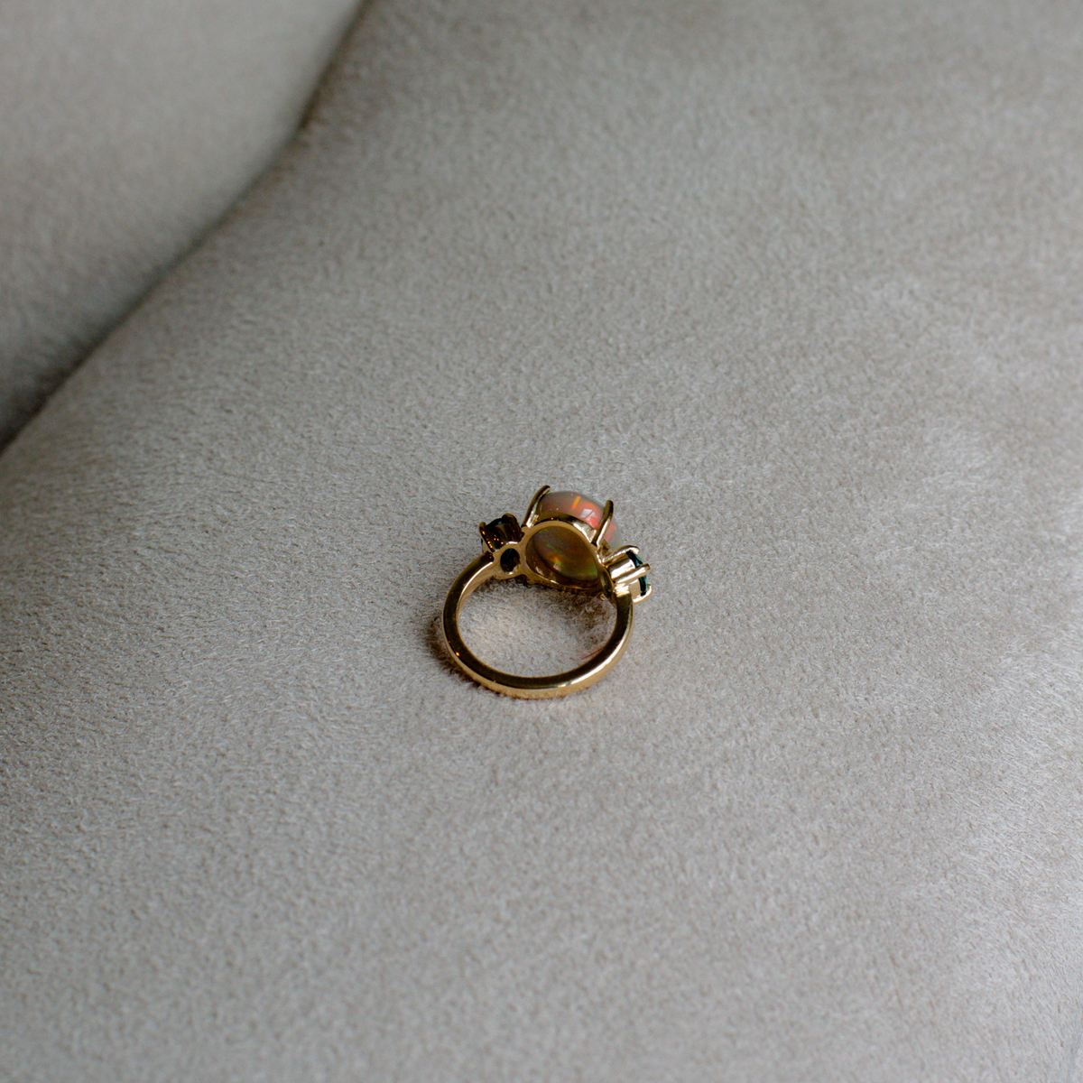 Back side of a gold ring with an opal center and indicolite stones, resting on a gray suede surface.