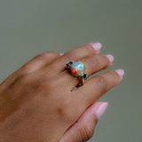 A gold ring featuring a round iridescent opal gemstone and small dark green stones on either side, shown worn on a hand with pink manicured nails against a solid gray background.