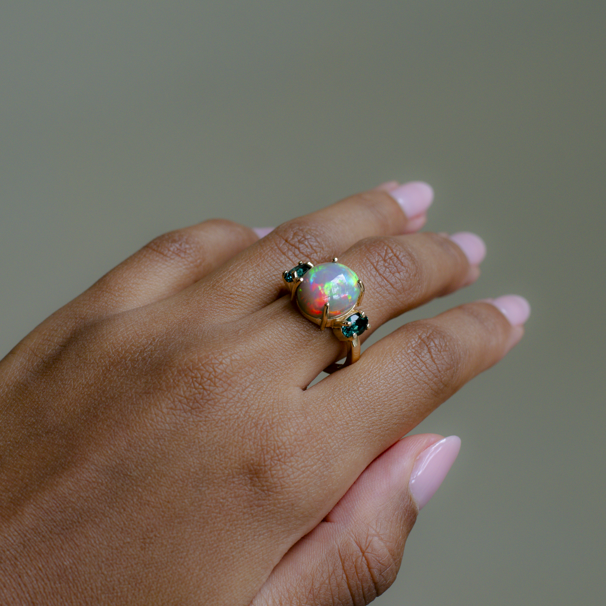 A gold ring featuring a round iridescent opal gemstone and small dark green stones on either side, shown worn on a hand with pink manicured nails against a solid gray background.
