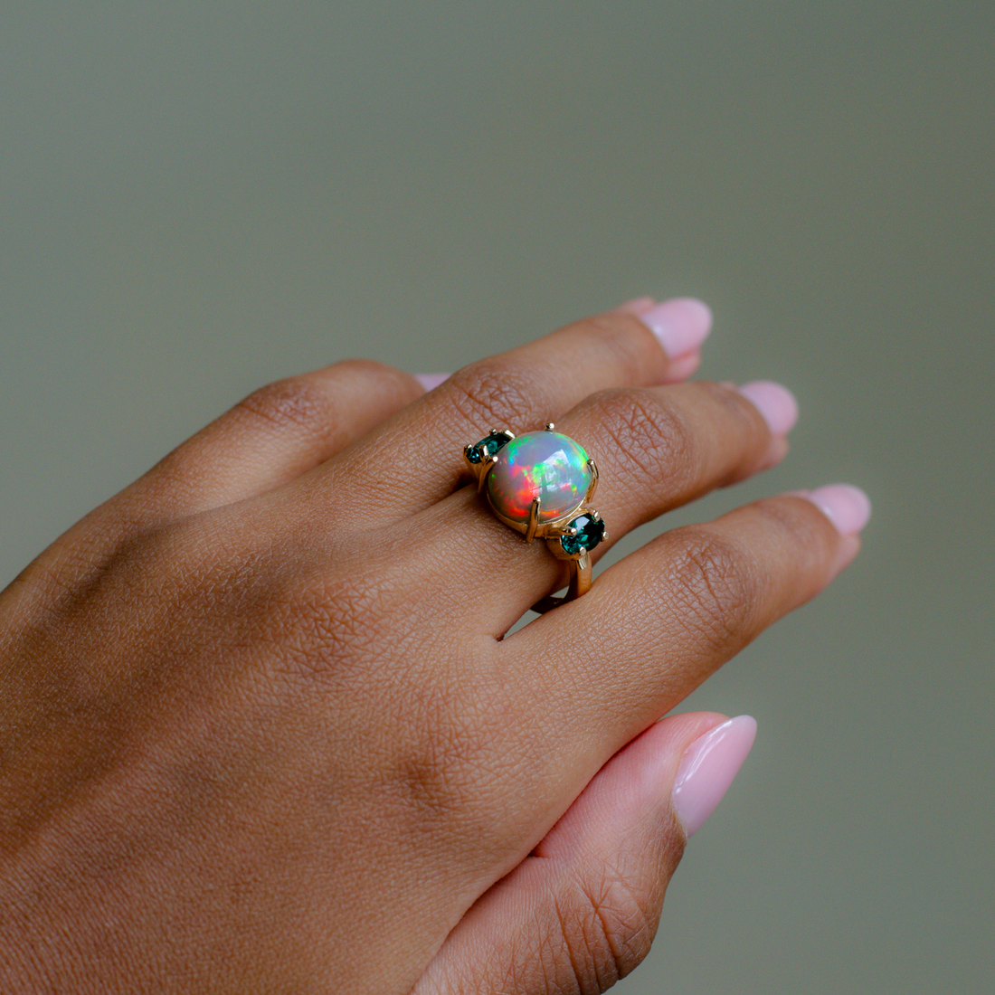 A gold ring featuring a round iridescent opal gemstone and small dark green stones on either side, shown worn on a hand with pink manicured nails against a solid gray background.