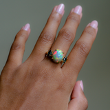 A close-up of a hand wearing a ring with a large round iridescent opal center stone and small dark stones on either side, set in a gold-toned band.