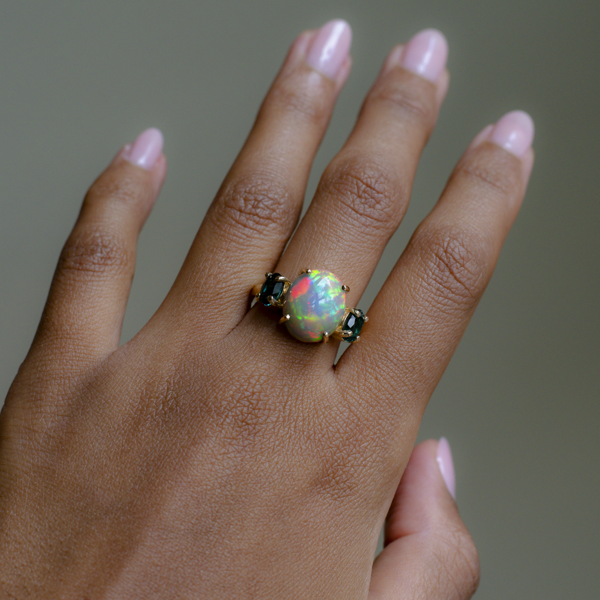 A close-up of a hand wearing a ring with a large round iridescent opal center stone and small dark stones on either side, set in a gold-toned band.