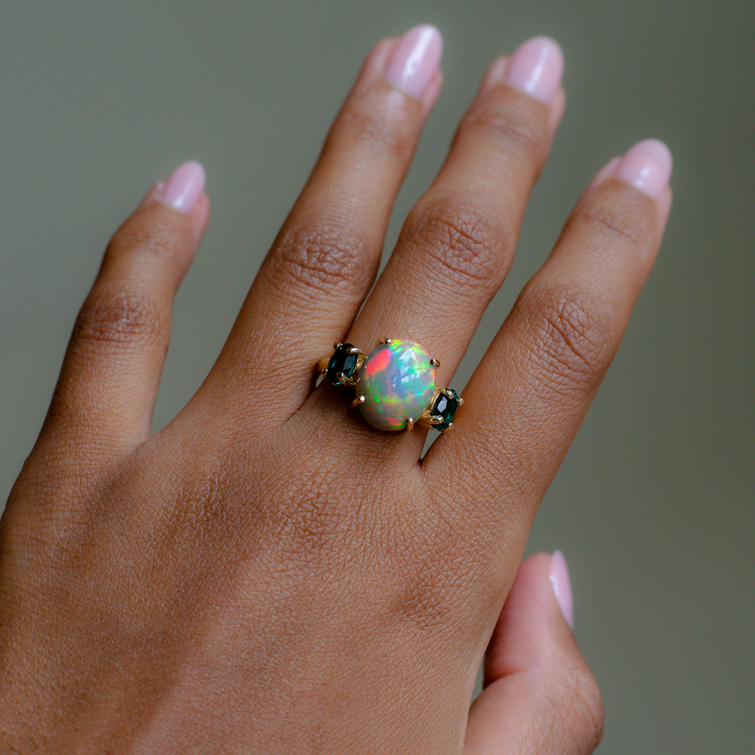 A close-up of a hand wearing a ring with a large round iridescent opal center stone and small dark stones on either side, set in a gold-toned band.
