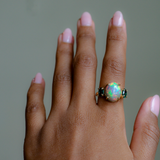 A close-up of a hand wearing a gold ring with a central iridescent opal gemstone and small dark green side stones, set against a soft gray background.
