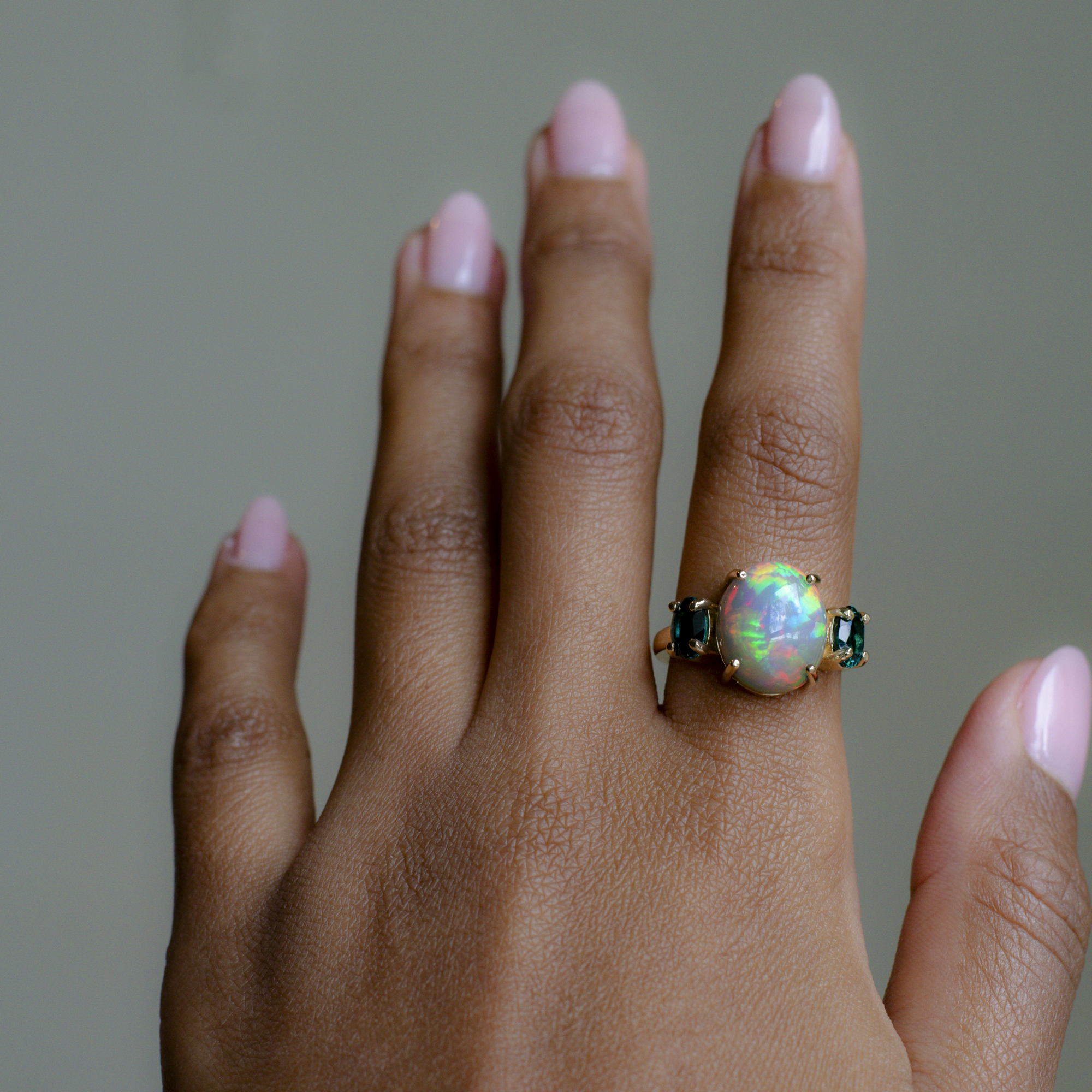A close-up of a hand wearing a gold ring with a central iridescent opal gemstone and small dark green side stones, set against a soft gray background.
