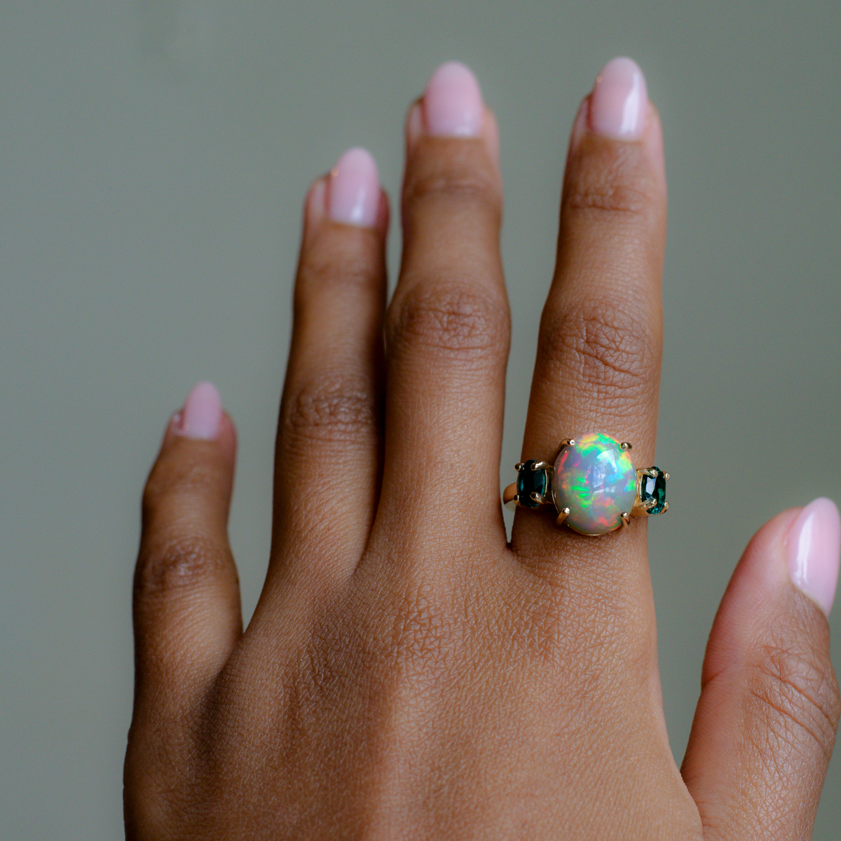 A close-up of a hand wearing a gold ring with a central iridescent opal gemstone and small dark green side stones, set against a soft gray background.
