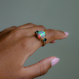 A gold ring featuring a large round iridescent opal center stone and two small dark green gemstones on either side, shown worn on a person's finger against a muted gray-green background.