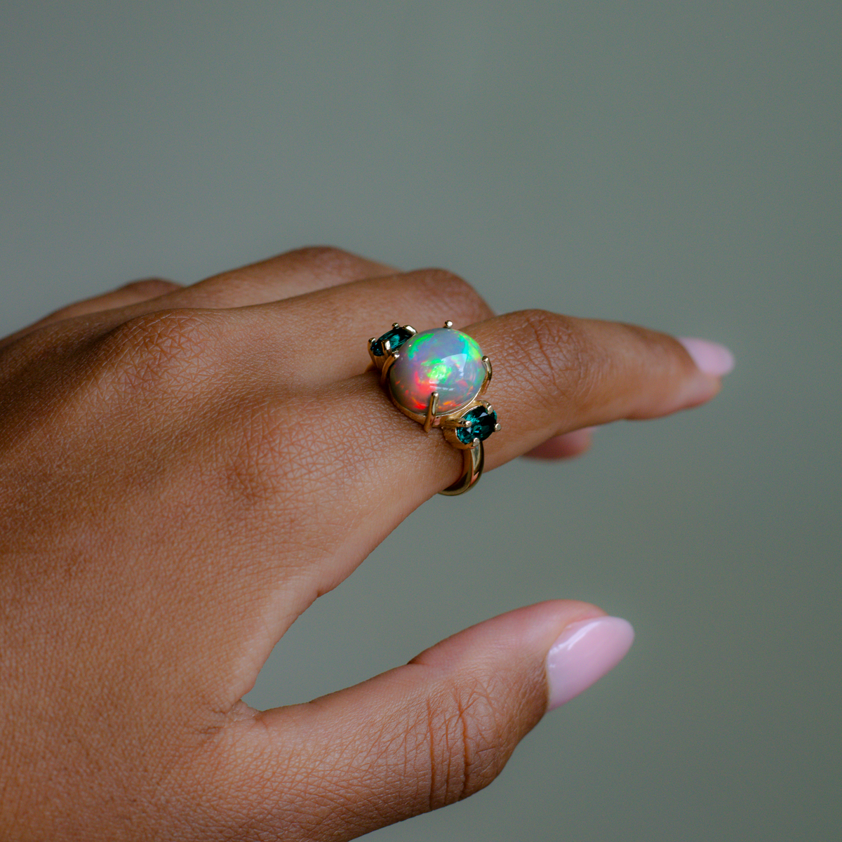 A gold ring featuring a large round iridescent opal center stone and two small dark green gemstones on either side, shown worn on a person's finger against a muted gray-green background.