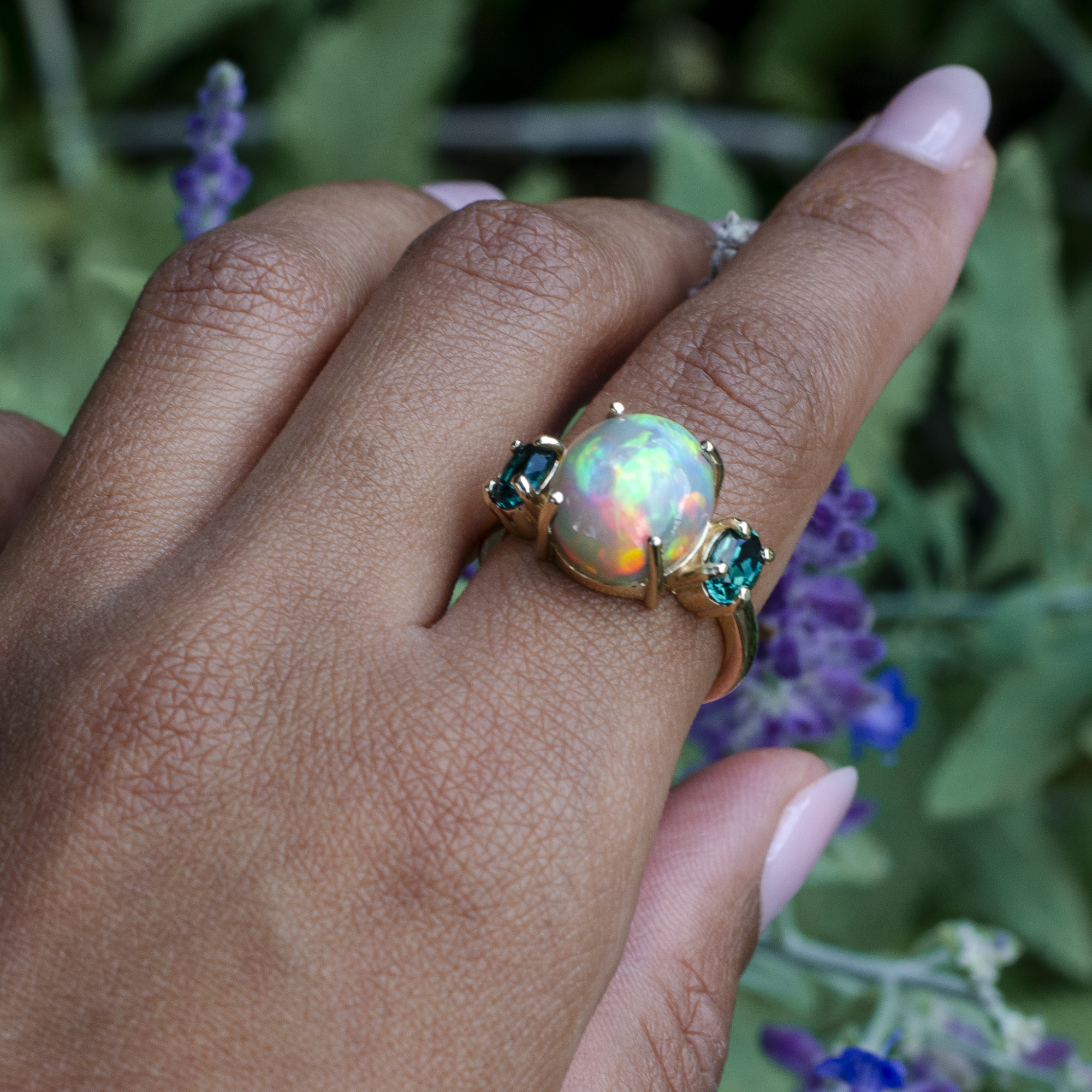 A gold ring featuring a large iridescent opal center stone and two green gemstones on either side, worn on a person's hand with blurred purple flowers in the background.