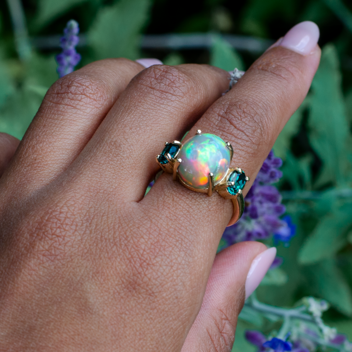 A gold ring featuring a large iridescent opal center stone and two green gemstones on either side, worn on a person's hand with blurred purple flowers in the background.