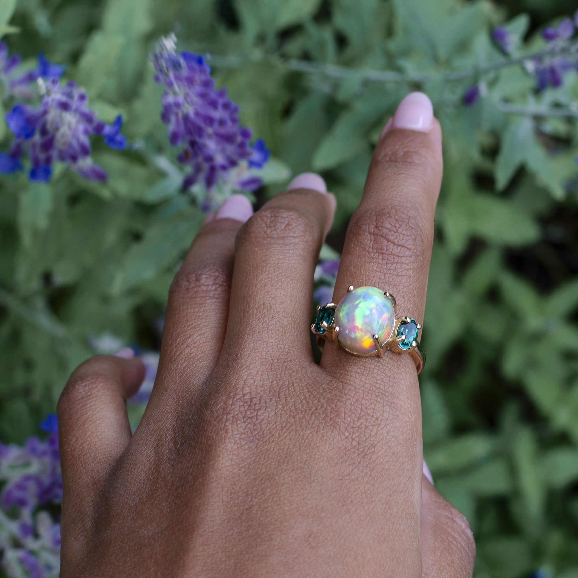 A gold ring featuring a large iridescent opal center stone and two small teal-blue stones on either side, worn on a person's ring finger with a blurred background of green leaves and purple flowers.
