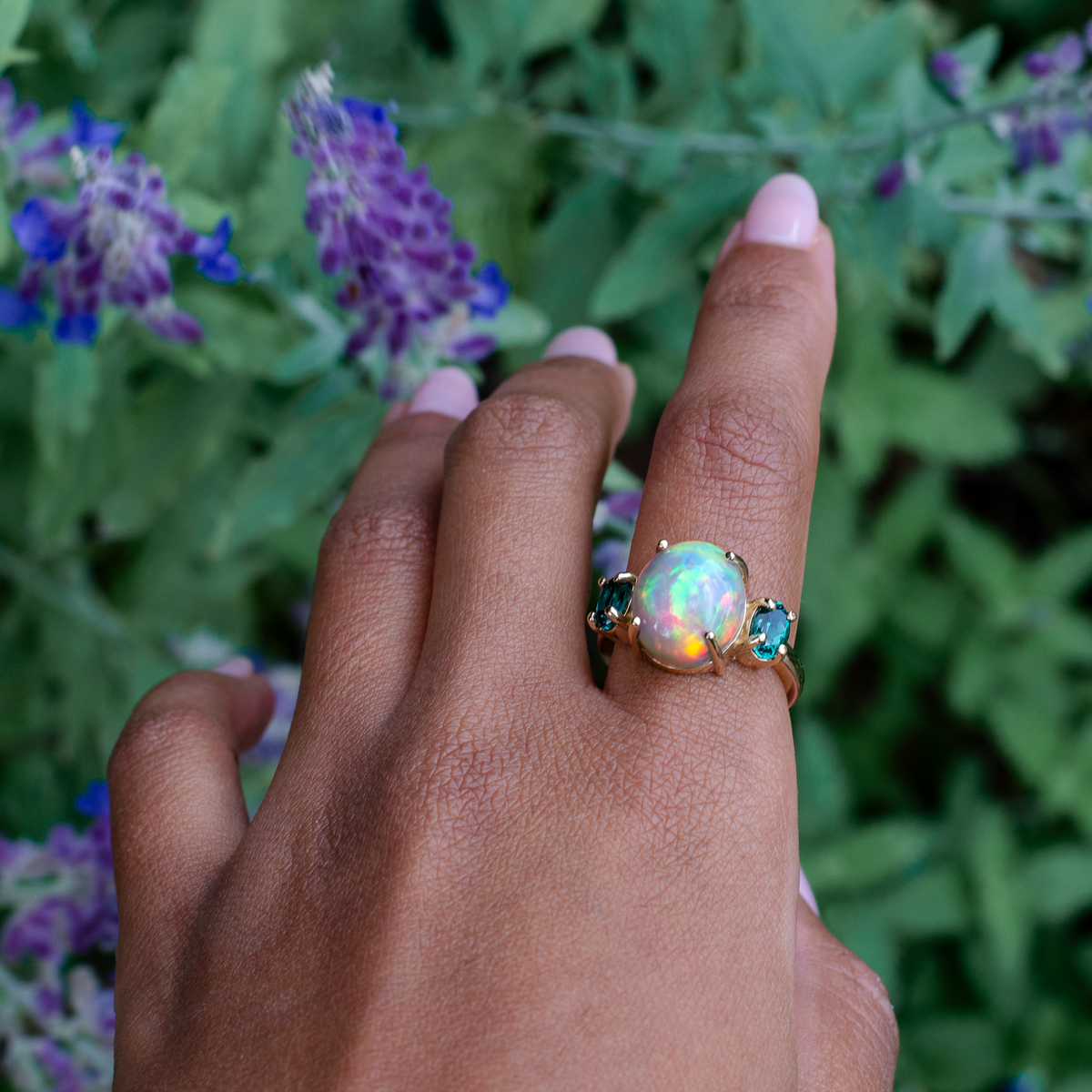 A gold ring featuring a large iridescent opal center stone and two small teal-blue stones on either side, worn on a person's ring finger with a blurred background of green leaves and purple flowers.