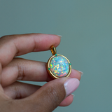 Hand holding a yellow gold pendant with a top quality Ethiopian opal and emerald accent stones. 