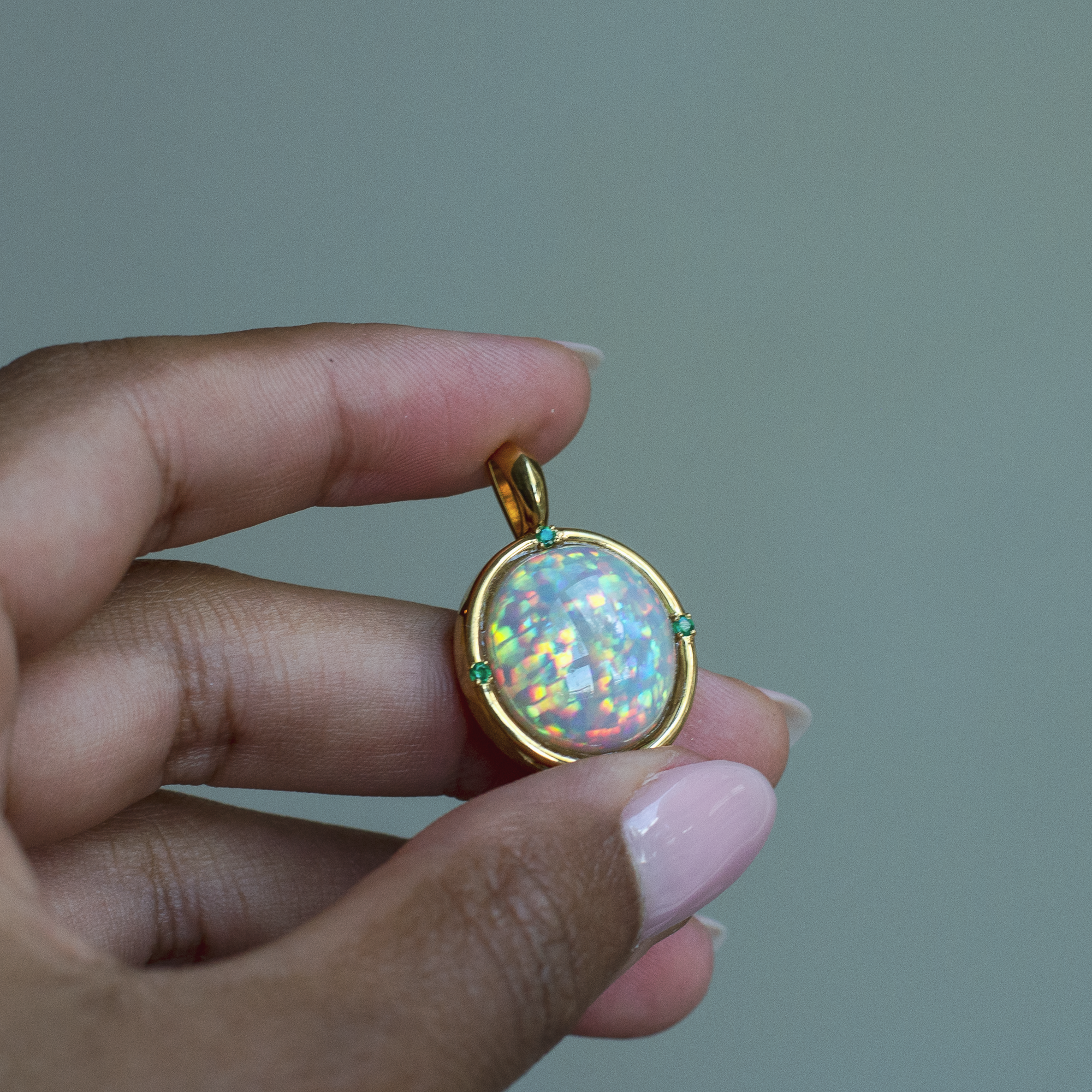 Hand holding a yellow gold pendant with a top quality Ethiopian opal and emerald accent stones. 