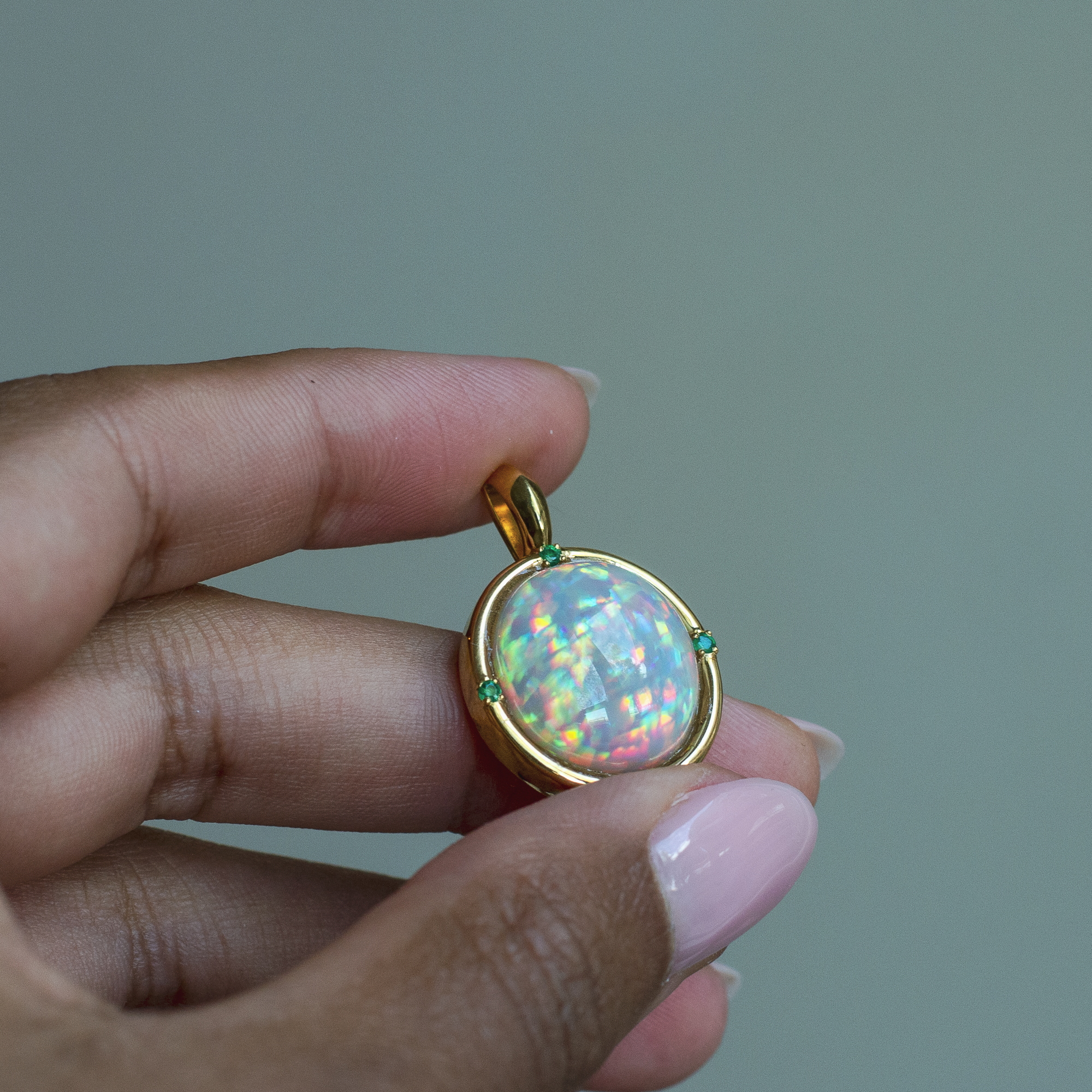 Hand holding a 14k yellow gold bezel pendant with a top quality oval Ethiopian opal and emerald accent stones. 