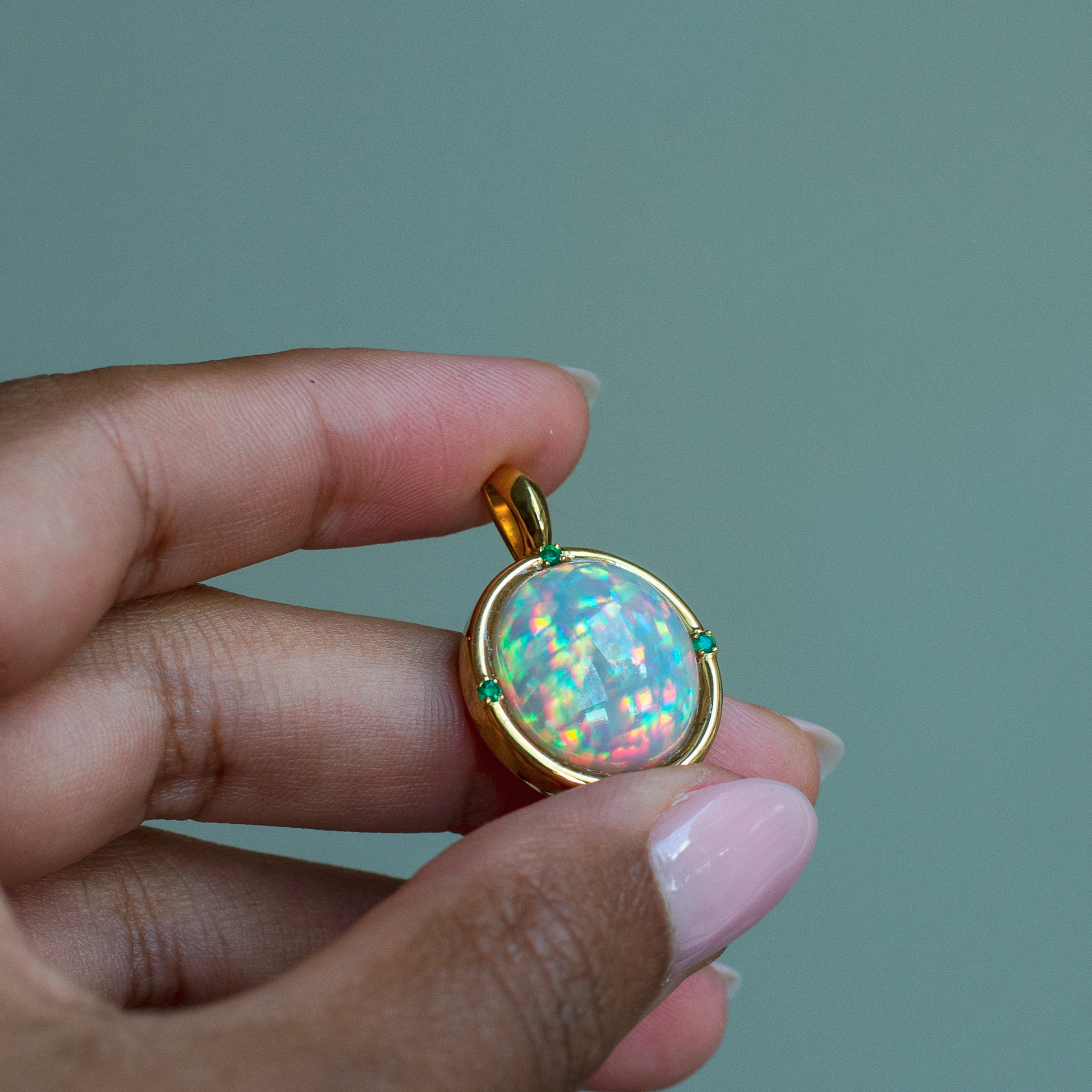 Hand holding a 14k yellow gold bezel pendant with a top quality oval Ethiopian opal and emerald accent stones. 