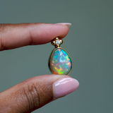 14k solid yellow gold Ethiopian Opal pendant with diamond accent and gold granulation beading flower. Highest quality, museum-grade AAA gemstone from Betami Gems. Held between fingers against a neutral background.