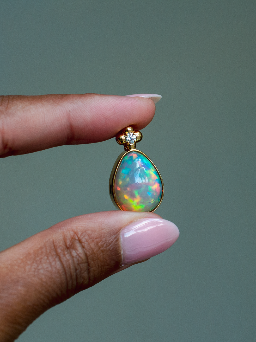 14k solid yellow gold Ethiopian Opal pendant with diamond accent and gold granulation beading flower. Highest quality, museum-grade AAA gemstone from Betami Gems. Held between fingers against a neutral background.