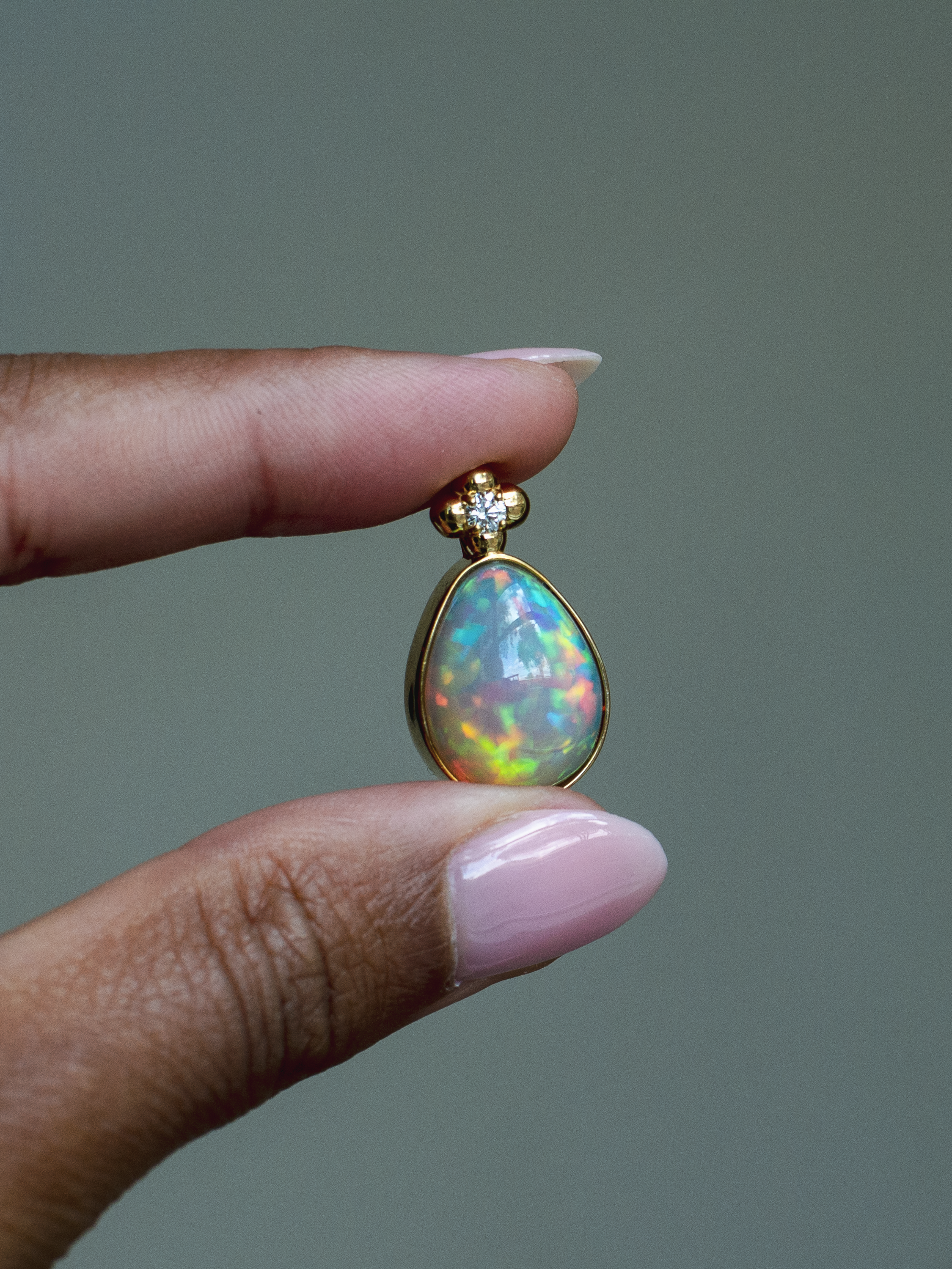 14k solid yellow gold Ethiopian Opal pendant with diamond accent and gold granulation beading flower. Highest quality, museum-grade AAA gemstone from Betami Gems. Held between fingers against a neutral background.