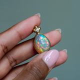14k solid yellow gold Ethiopian Opal pendant with diamond accent and gold granulation beading flower. Held between fingers against a neutral background.