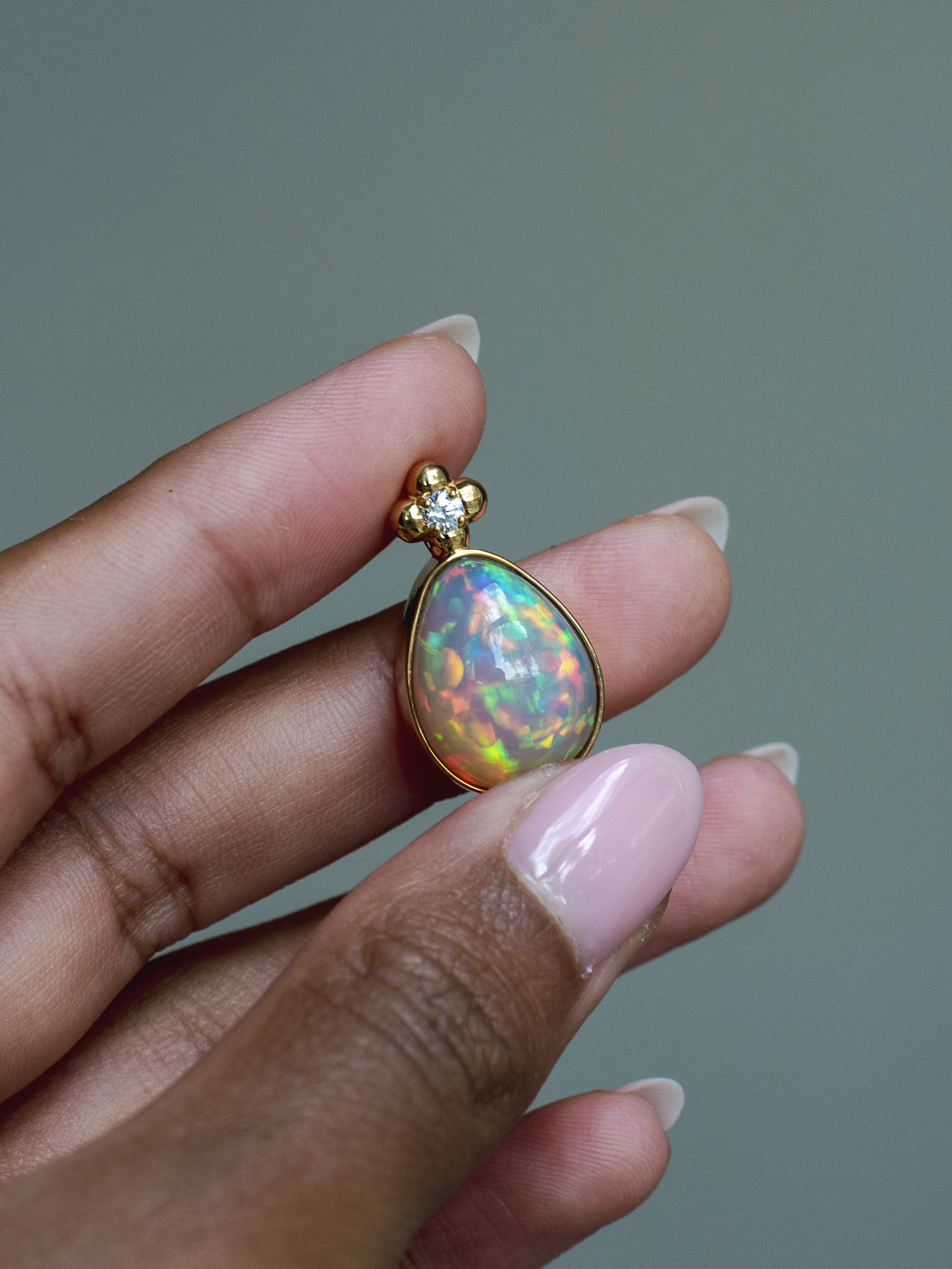14k solid yellow gold Ethiopian Opal pendant with diamond accent and gold granulation beading flower. Held between fingers against a neutral background.