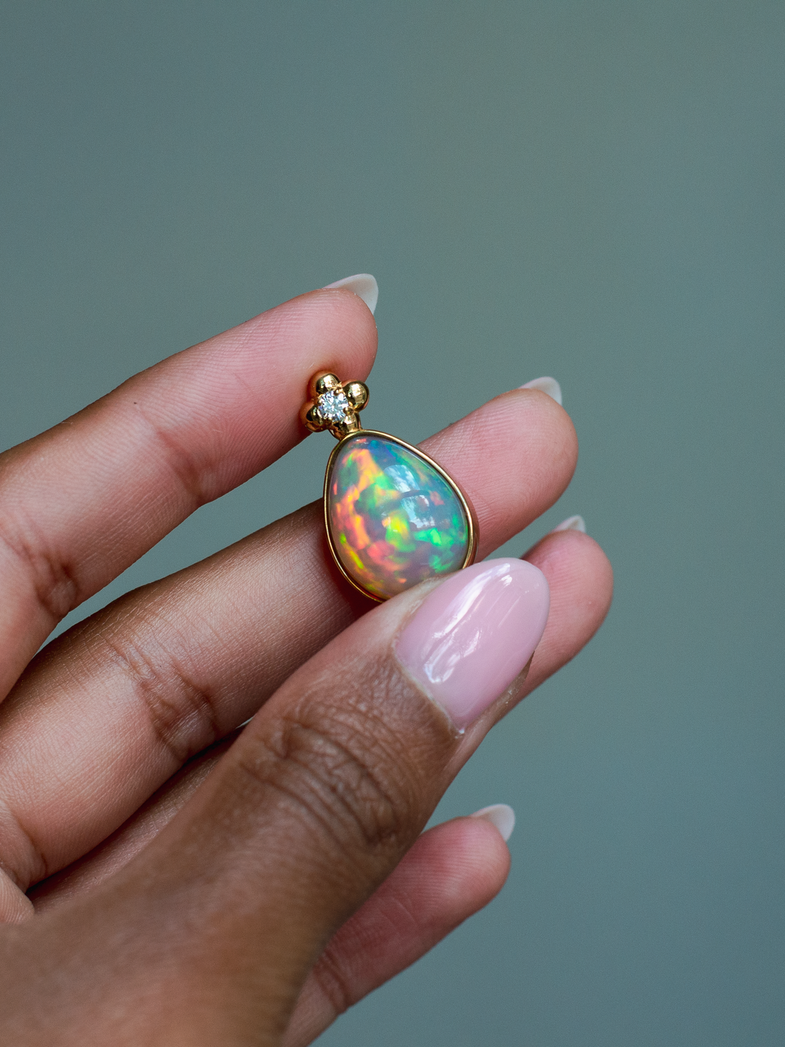 14k solid yellow gold Ethiopian Opal pendant with diamond accent and gold granulation beading flower. Held between fingers against a neutral background.
