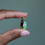 Side view of 14k solid yellow gold Ethiopian Opal bezel set pendant with diamond accent and gold granulation beading flower. Held between fingers against a neutral background.