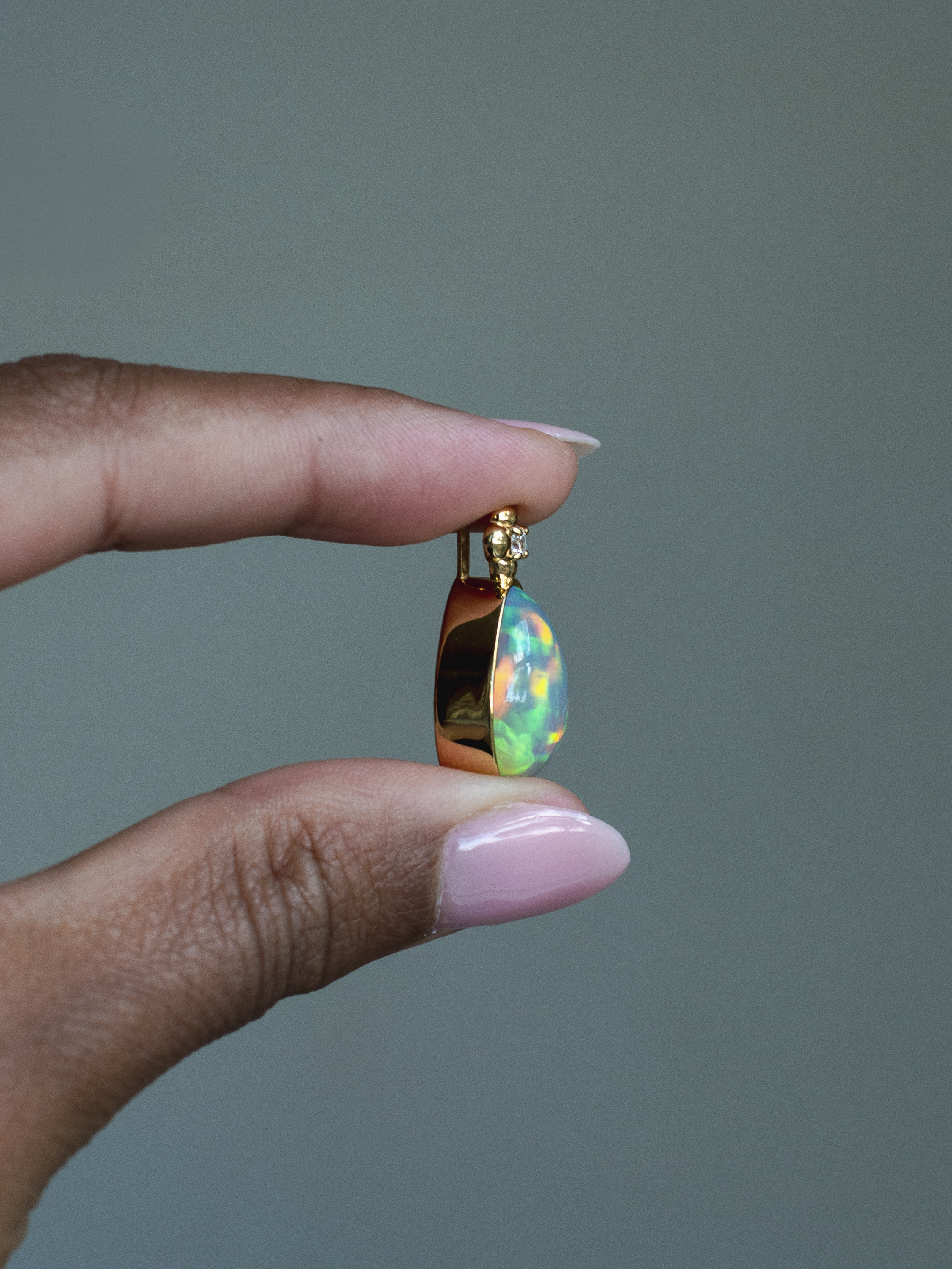 Side view of 14k solid yellow gold Ethiopian Opal bezel set pendant with diamond accent and gold granulation beading flower. Held between fingers against a neutral background.