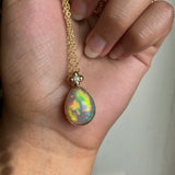 14k solid yellow gold Ethiopian Opal bezel set necklace with chain and diamond accent and gold granulation beading flower. Strong play of fire with lots of yellow and orange. Held by a hand against a white wall.