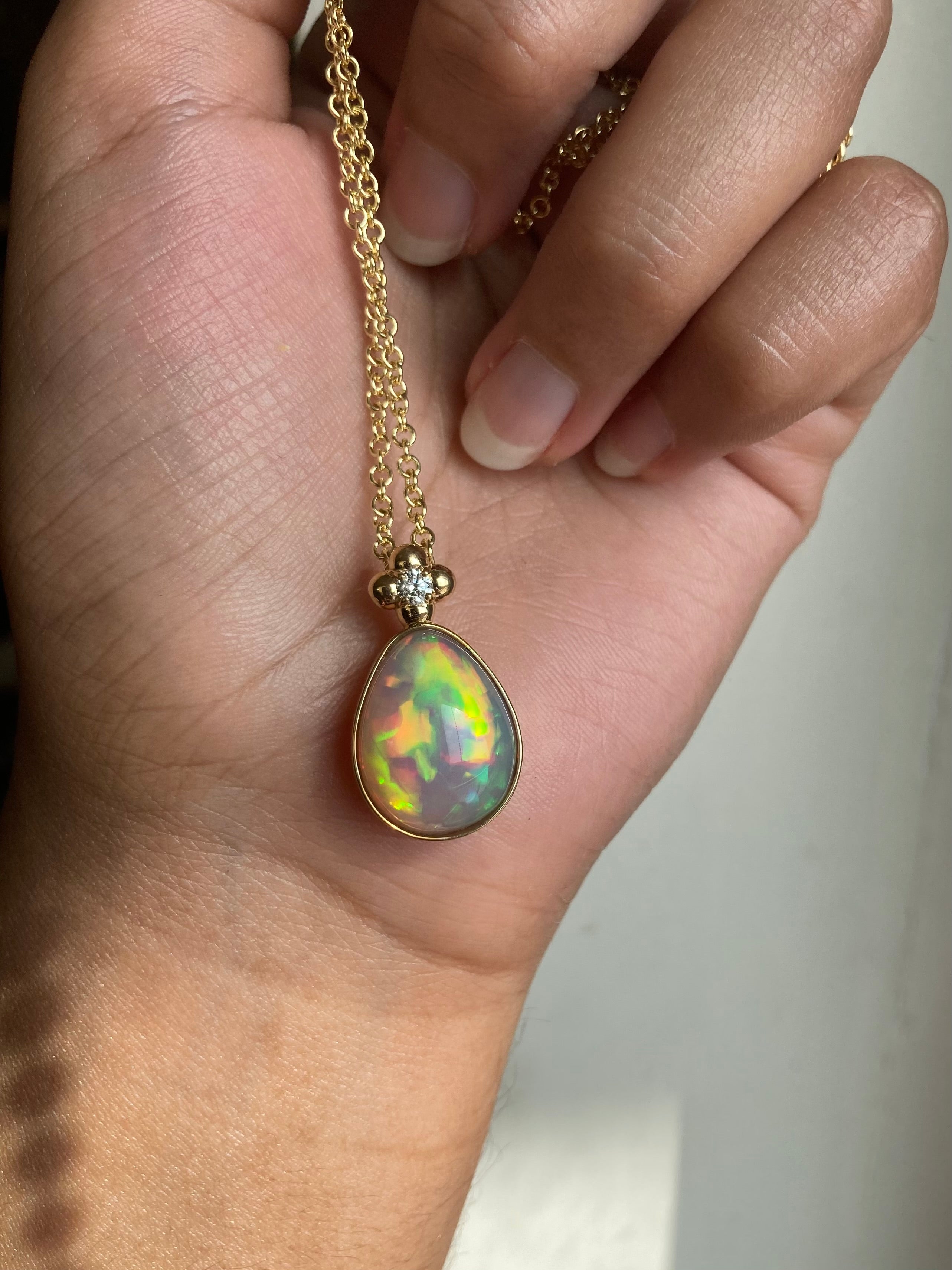 14k solid yellow gold Ethiopian Opal bezel set necklace with chain and diamond accent and gold granulation beading flower. Strong play of fire with lots of yellow and orange. Held by a hand against a white wall.