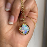 A hand holding a solid 14k yellow gold necklace with a multicolored opal gemstone held in a bezel setting the shape of a four leaf clover.