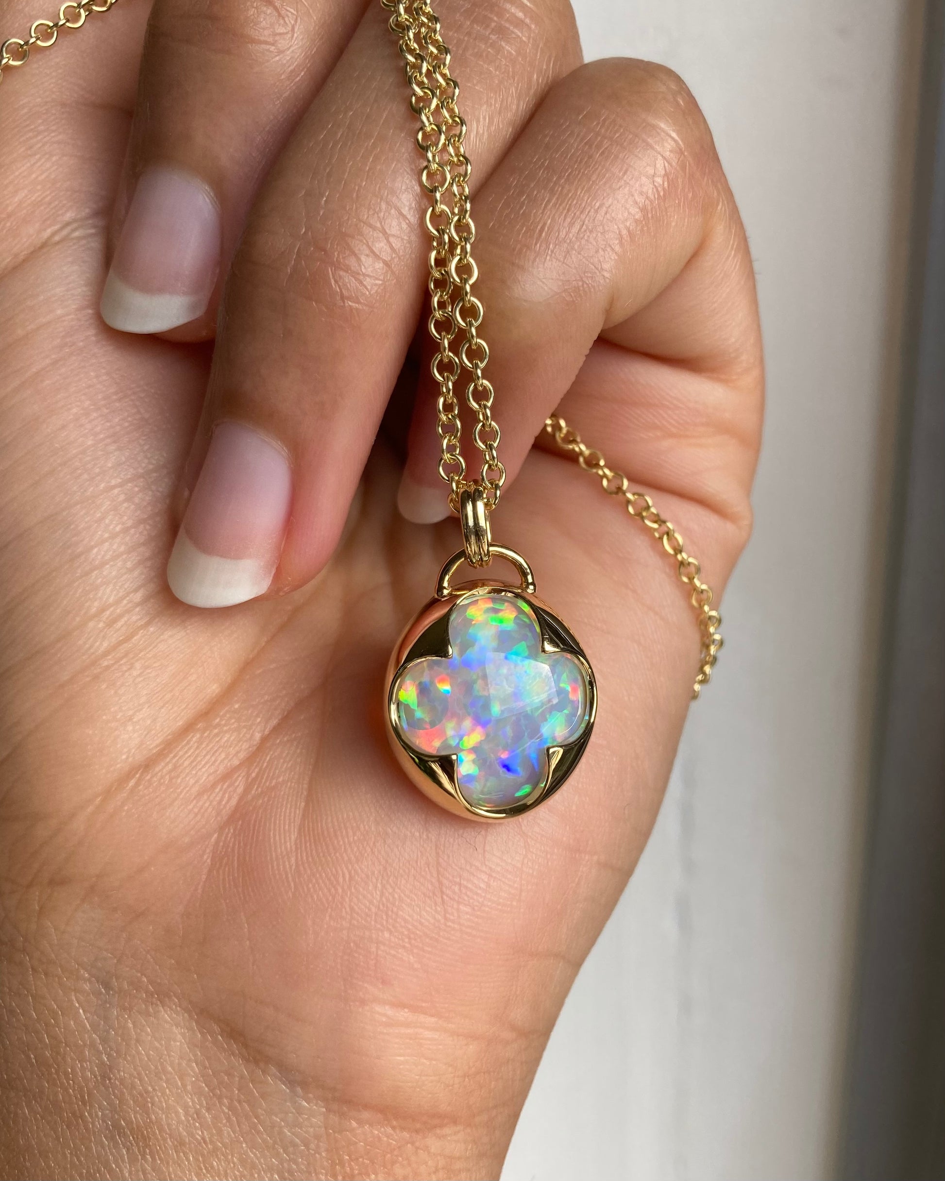A hand holding a solid 14k yellow gold necklace with a multicolored opal gemstone held in a bezel setting the shape of a four leaf clover.