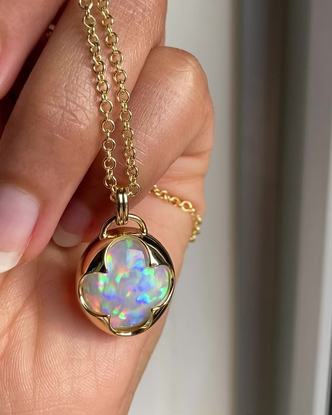 A hand holding a solid 14k yellow gold pendant with a multicolored opal gemstone held in a bezel setting the shape of a four leaf clover, with a gold chain.